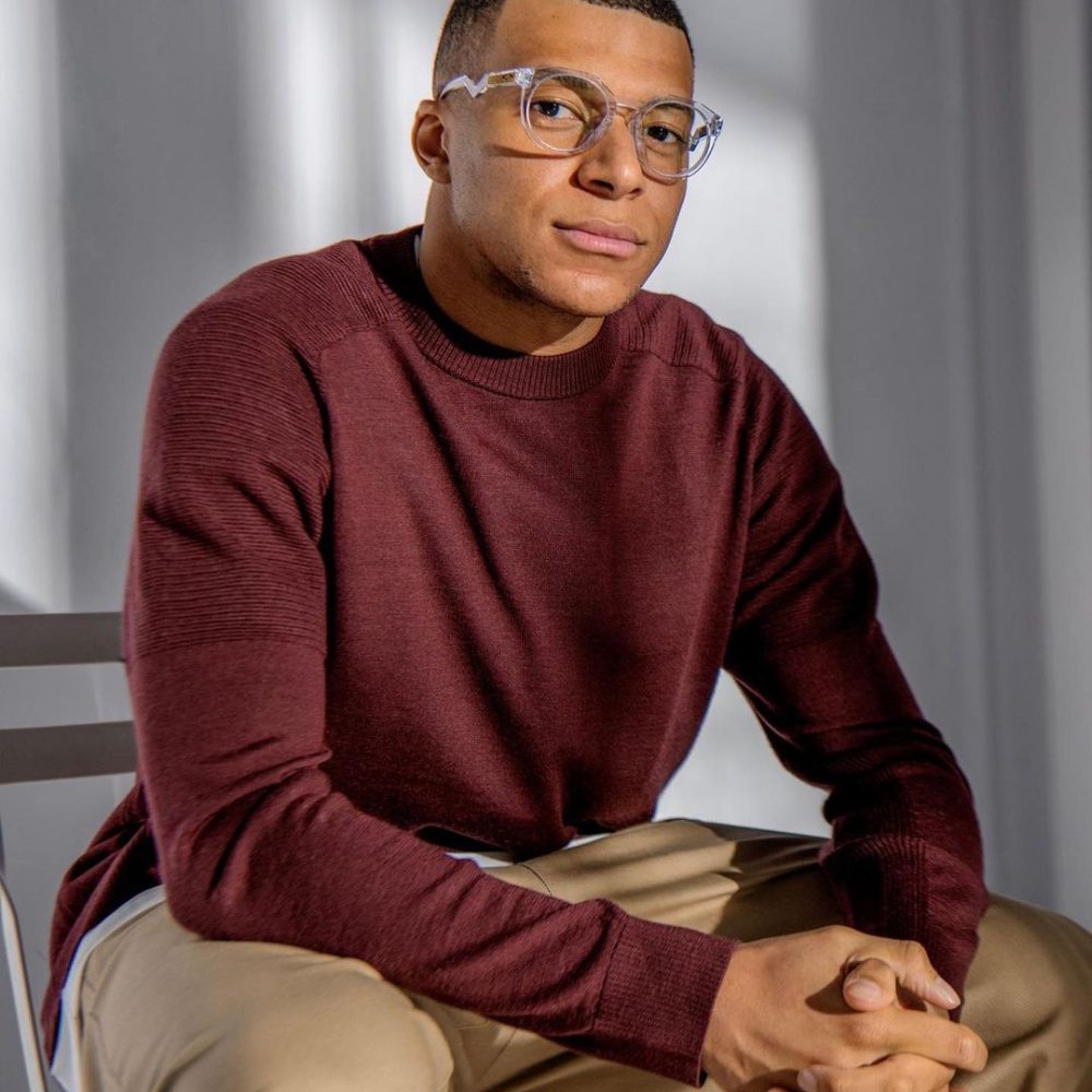 Kylian Mbappé sitzt mit einem weinroten Pullover und einer Nerd-Brille auf einem weißen Stuhl und schaut ernst in die Kamera.